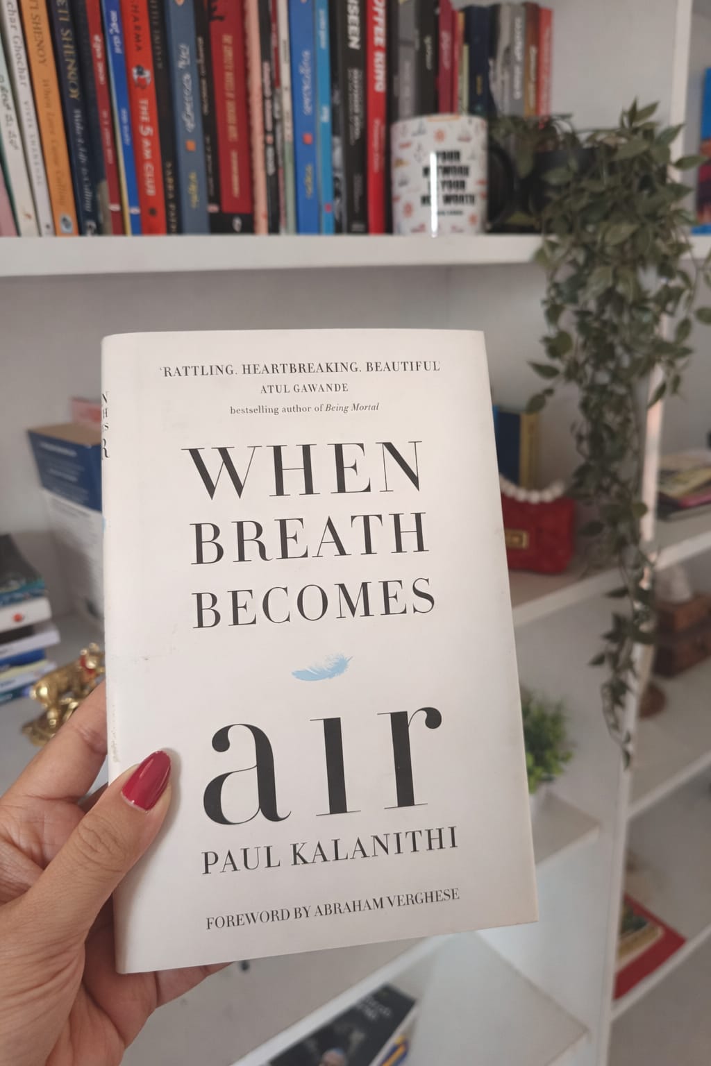 Hand holding the book When Breath Becomes Air by Paul Kalanithi in front of a bookshelf.